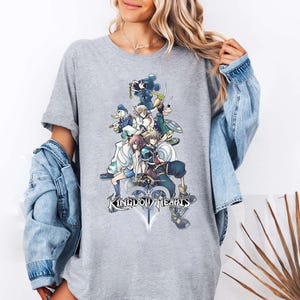 Puede incluir: Una camiseta gris con un gr&aacute;fico colorido de personajes del videojuego "Kingdom Hearts". El dise&ntilde;o incluye m&uacute;ltiples figuras, una espada y el t&iacute;tulo del juego. La camiseta es una prenda casual.