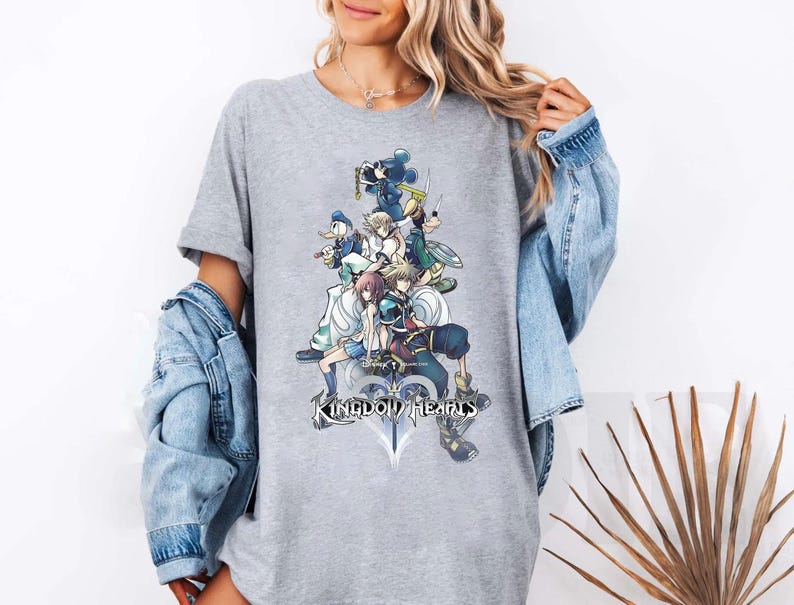 Puede incluir: Una camiseta gris con un gr&aacute;fico colorido de personajes del videojuego "Kingdom Hearts". El dise&ntilde;o incluye m&uacute;ltiples figuras, una espada y el t&iacute;tulo del juego. La camiseta es una prenda casual.