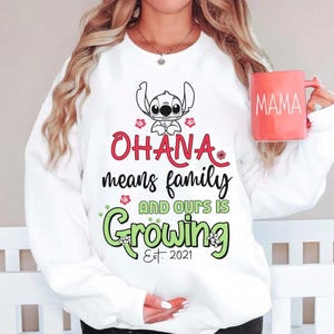 May include: White sweatshirt with the text "OHANA means family AND OURS IS Growing Est. 2021" and a cartoon character illustration. The sweatshirt also features a red mug with the word "MAMA" on it.