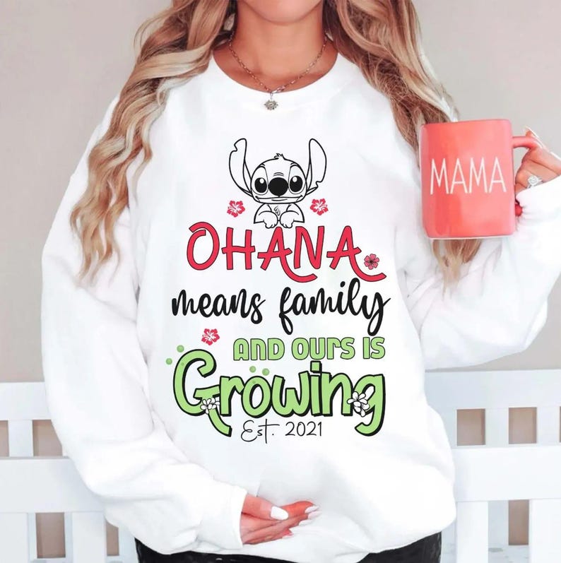 May include: White sweatshirt with the text "OHANA means family AND OURS IS Growing Est. 2021" and a cartoon character illustration. The sweatshirt also features a red mug with the word "MAMA" on it.
