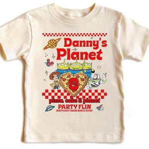 Pu&ograve; includere: T-shirt color crema con un design grafico colorato. Il design include il testo "Danny's Planet" in rosso, personaggi dei cartoni animati e un otto a forma di pizza. La maglietta ha anche il testo "Pizza, Cake & Planet PARTY FLIN BIRTHDAY CREW SINCE 2016!"