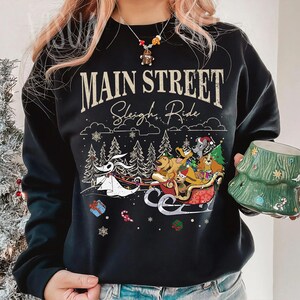 May include: Black sweatshirt with the text "Main Street Sleigh Ride" and a graphic of a sleigh pulled by dogs and a character from The Nightmare Before Christmas. The design includes Christmas trees, snowflakes, and presents.