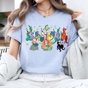 May include: Light blue t-shirt featuring the name "SANDRA" in colorful, cartoon-style lettering. Each letter is decorated with aquatic characters and elements, including mermaids, fish, and seaweed.