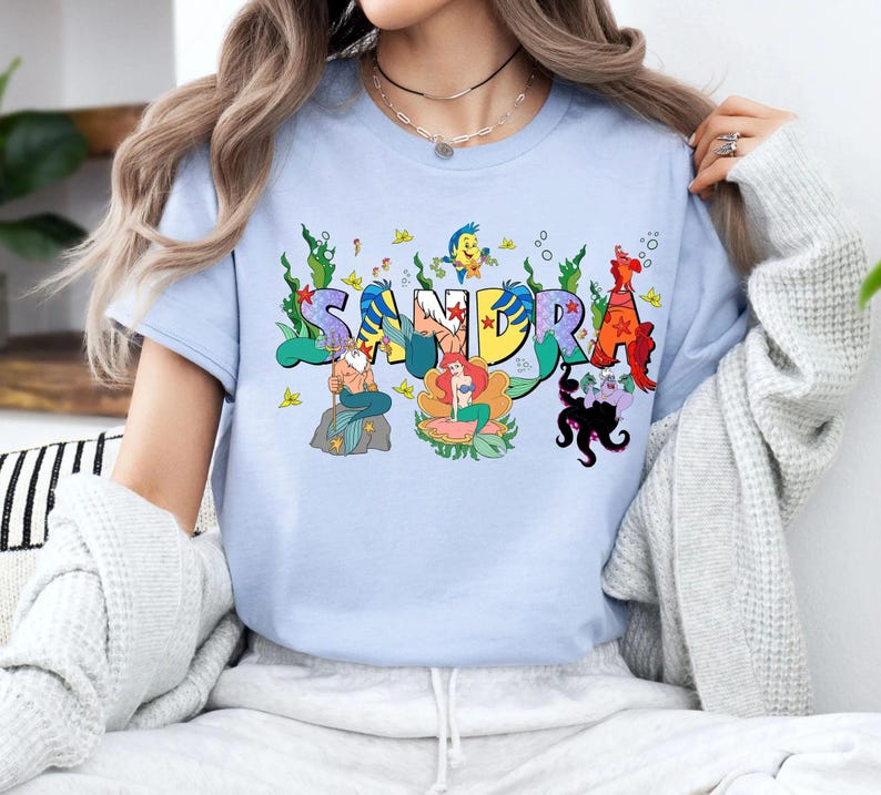May include: Light blue t-shirt featuring the name "SANDRA" in colorful, cartoon-style lettering. Each letter is decorated with aquatic characters and elements, including mermaids, fish, and seaweed.