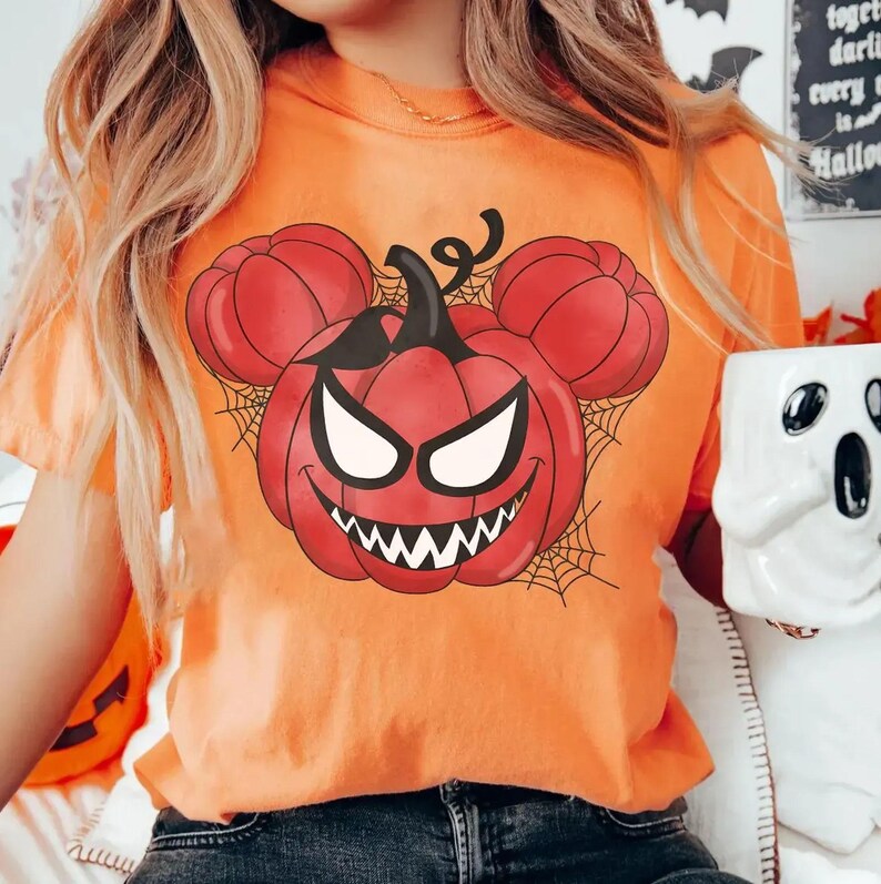May include: An orange t-shirt featuring a graphic of a red pumpkin with a sinister face and mouse-ear-shaped pumpkin side accents. The design includes black spiderweb patterns behind the pumpkin, creating a festive Halloween-themed illustration.