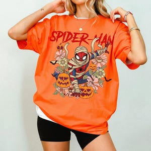 K&ouml;nnte beinhalten: Orangefarbenes T-Shirt mit dem roten Schriftzug "SPIDER-MAN". Die Grafik zeigt Spider-Man als Mumie, umgeben von K&uuml;rbissen und Blumen. Das Shirt hat ein wei&szlig;es Unterhemd und schwarze Shorts.