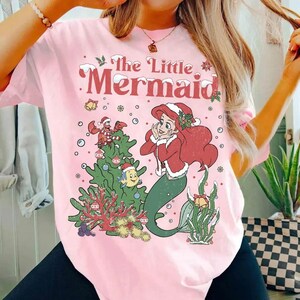 K&ouml;nnte beinhalten: Hellrosa T-Shirt mit einem weihnachtlichen Motiv von Arielle, der Meerjungfrau. Das Design umfasst einen Weihnachtsbaum, Schneeflocken und den Text "The Little Mermaid" in Rot.