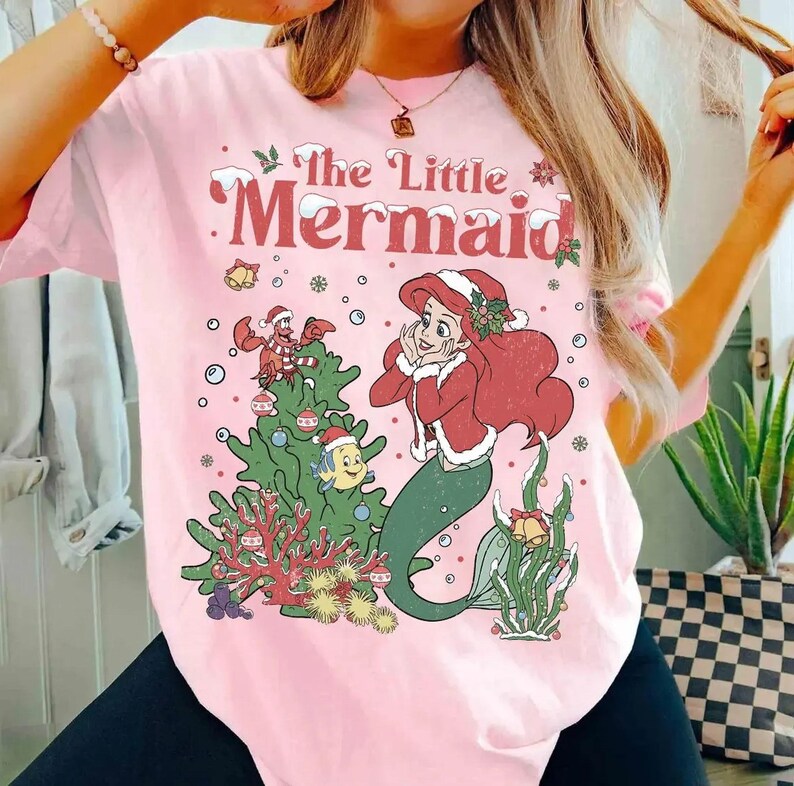 K&ouml;nnte beinhalten: Hellrosa T-Shirt mit einem weihnachtlichen Motiv von Arielle, der Meerjungfrau. Das Design umfasst einen Weihnachtsbaum, Schneeflocken und den Text "The Little Mermaid" in Rot.