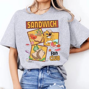 May include: A light gray t-shirt featuring a colorful graphic design. The design includes the word "SANDWICH" in a brown rectangular box above three square illustrations of sandwiches. Below the illustrations is the text "FAN CLUB".