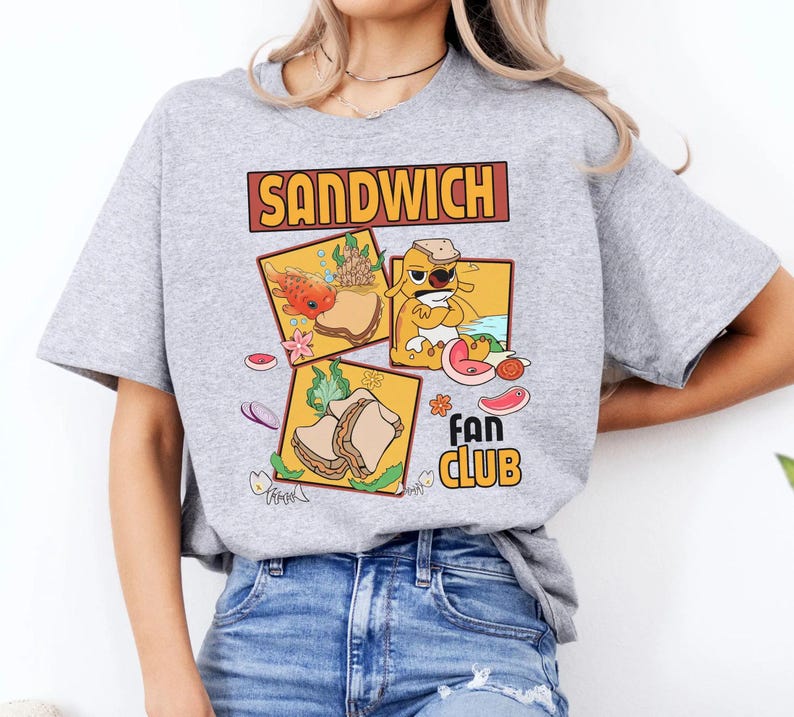May include: A light gray t-shirt featuring a colorful graphic design. The design includes the word "SANDWICH" in a brown rectangular box above three square illustrations of sandwiches. Below the illustrations is the text "FAN CLUB".