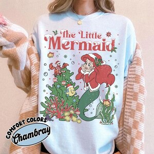K&ouml;nnte beinhalten: Hellblaues Sweatshirt mit einer festlichen Illustration von Arielle, der Meerjungfrau. Das Design zeigt Arielle mit Weihnachtsm&uuml;tze und roter Jacke, einen Weihnachtsbaum und den Text "The Little Mermaid" in Rot. Auf dem Sweatshirt steht "Comfort Colors Chambray".