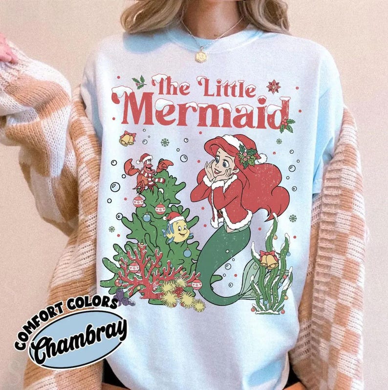 K&ouml;nnte beinhalten: Hellblaues Sweatshirt mit einer festlichen Illustration von Arielle, der Meerjungfrau. Das Design zeigt Arielle mit Weihnachtsm&uuml;tze und roter Jacke, einen Weihnachtsbaum und den Text "The Little Mermaid" in Rot. Auf dem Sweatshirt steht "Comfort Colors Chambray".