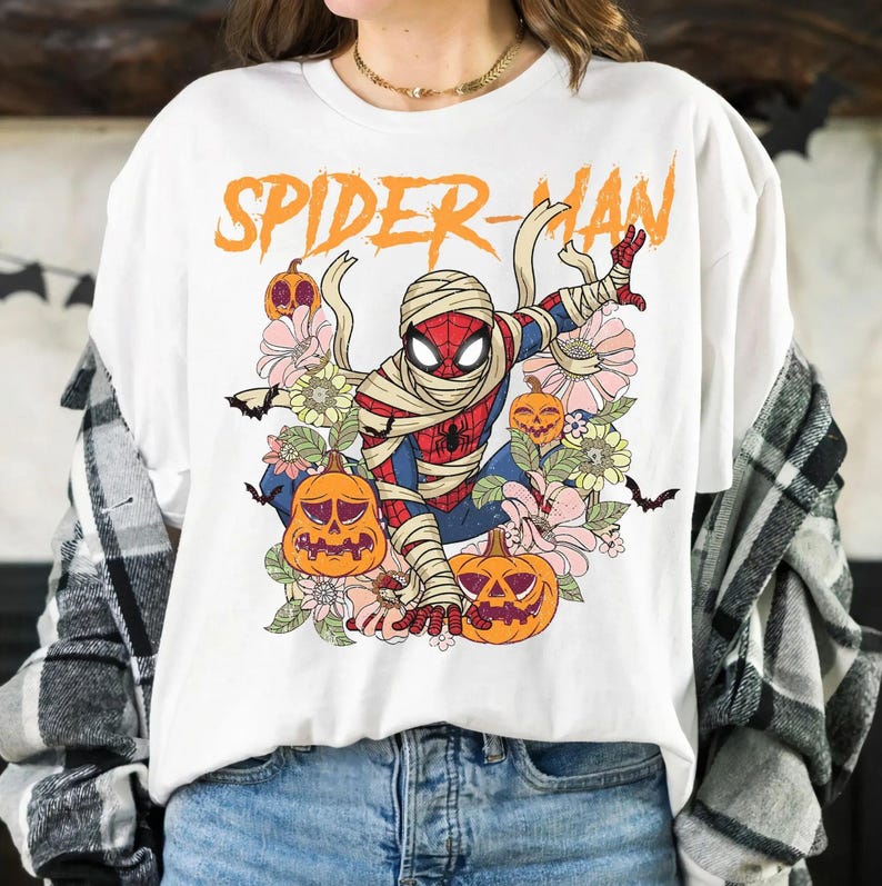 K&ouml;nnte beinhalten: Wei&szlig;es T-Shirt mit einer Grafik von Spider-Man als Mumie, umgeben von K&uuml;rbissen und Blumen. Der Text "SPIDER-MAN" ist orange. Das Design hat ein Vintage-Halloween-Thema.