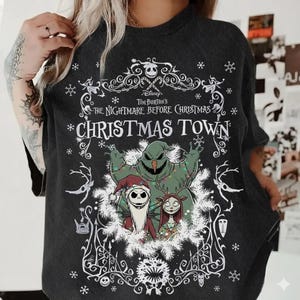 May include: Dark gray t-shirt featuring a graphic from Tim Burton's "The Nightmare Before Christmas." The design includes Jack Skellington, Sally, and Oogie Boogie, with the words "Christmas Town" in a decorative font. The shirt has a crew neck and short sleeves.