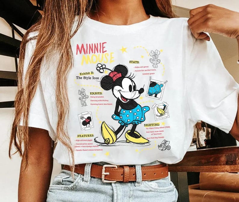 以下が含まれることがあります： ミニーマウスのグラフィックがプリントされた白いTシャツ。デザインには、水玉模様の青いドレス、黄色の靴、赤いリボンを着たミニーが含まれています。Tシャツのテキストは「ミニーマウス」と「Exhibit B: The Style Icon」です。