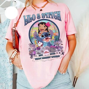 Może przedstawiać: R&oacute;żowa koszulka z grafiką Lilo & Stitch. Projekt zawiera niebieskie słowa "Lilo & Stitch", wizerunki postaci oraz tekst "Est 2002 Walt Disney World". Koszulka ma kr&oacute;tkie rękawy.