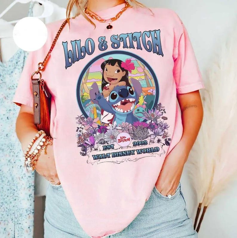 Może przedstawiać: R&oacute;żowa koszulka z grafiką Lilo & Stitch. Projekt zawiera niebieskie słowa "Lilo & Stitch", wizerunki postaci oraz tekst "Est 2002 Walt Disney World". Koszulka ma kr&oacute;tkie rękawy.