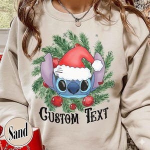 Puede incluir: Una sudadera color arena con un dise&ntilde;o navide&ntilde;o. El gr&aacute;fico presenta un personaje de dibujos animados azul con un gorro de Pap&aacute; Noel, rodeado de ramas de pino verdes y adornos rojos. El texto "CUSTOM TEXT" est&aacute; impreso en negro.
