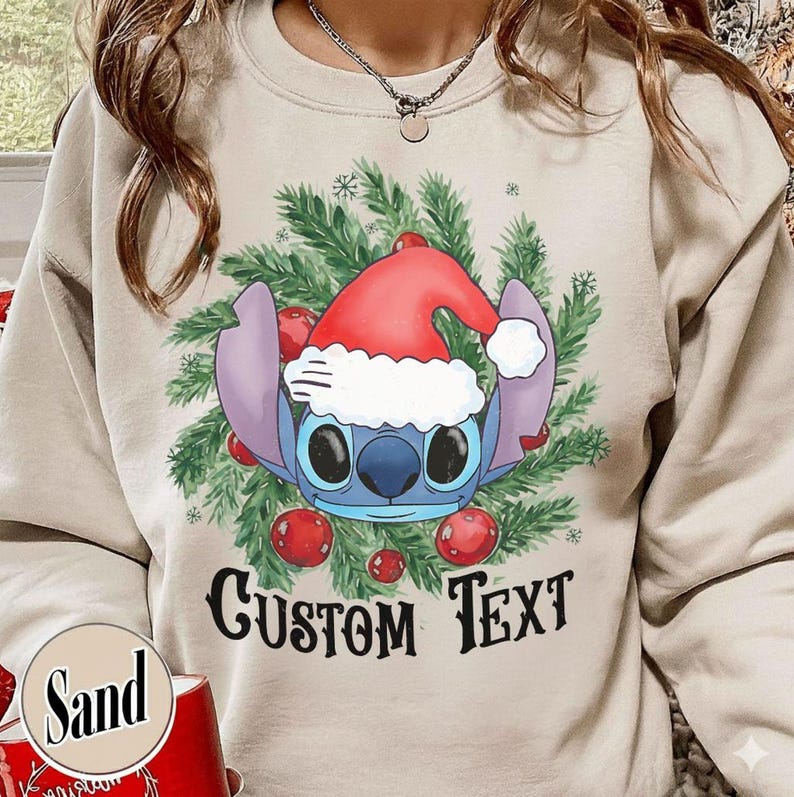 Puede incluir: Una sudadera color arena con un dise&ntilde;o navide&ntilde;o. El gr&aacute;fico presenta un personaje de dibujos animados azul con un gorro de Pap&aacute; Noel, rodeado de ramas de pino verdes y adornos rojos. El texto "CUSTOM TEXT" est&aacute; impreso en negro.