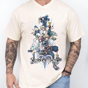 Puede incluir: Camiseta crema con cuello en V con un gr&aacute;fico de personajes de Kingdom Hearts, incluyendo Mickey Mouse y Donald Duck. El dise&ntilde;o presenta el logo del juego en la parte inferior. La camiseta es de color crema claro.