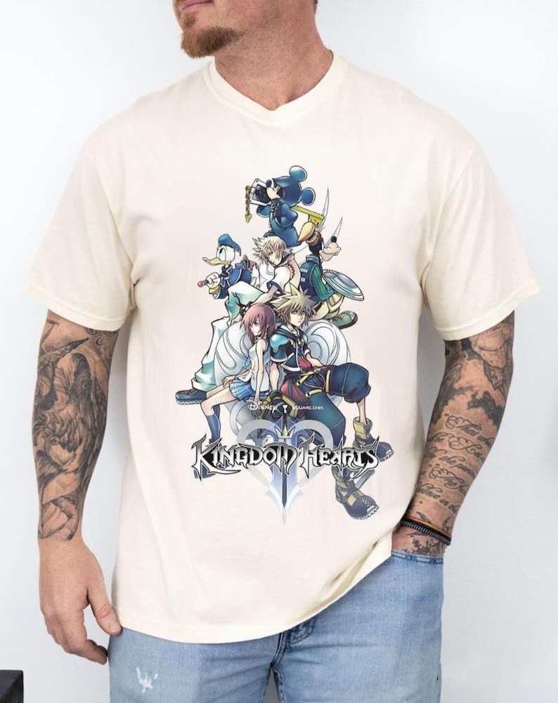 Puede incluir: Camiseta crema con cuello en V con un gr&aacute;fico de personajes de Kingdom Hearts, incluyendo Mickey Mouse y Donald Duck. El dise&ntilde;o presenta el logo del juego en la parte inferior. La camiseta es de color crema claro.