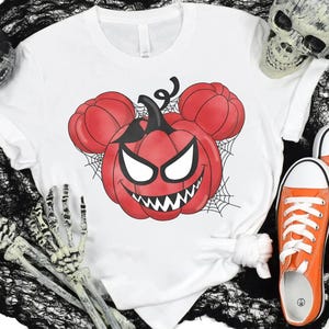 May include: A white short-sleeve t-shirt featuring a graphic of a red pumpkin with a jagged, toothy grin and white eyes, shaped like mouse ears, surrounded by black spiderwebs. The design is centered on the chest of the garment.
