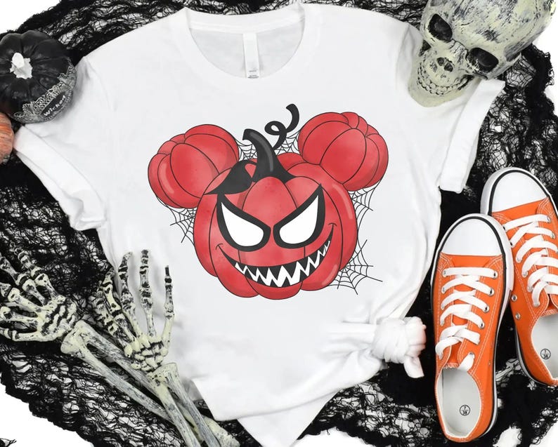 May include: A white short-sleeve t-shirt featuring a graphic of a red pumpkin with a jagged, toothy grin and white eyes, shaped like mouse ears, surrounded by black spiderwebs. The design is centered on the chest of the garment.