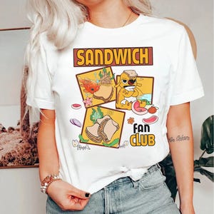 May include: White t-shirt featuring a "Sandwich Fan Club" design. The graphic includes cartoon illustrations of sandwiches and a dog character. The text "SANDWICH" is in a brown rectangular box.