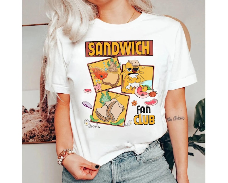 May include: White t-shirt featuring a "Sandwich Fan Club" design. The graphic includes cartoon illustrations of sandwiches and a dog character. The text "SANDWICH" is in a brown rectangular box.