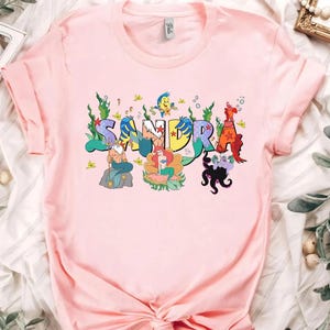 May include: A light pink t-shirt featuring the name "SANDRA" in colorful, whimsical lettering. Each letter is decorated with characters from a popular animated movie, including a mermaid, a crab, and a sea witch. The shirt is made of soft material.