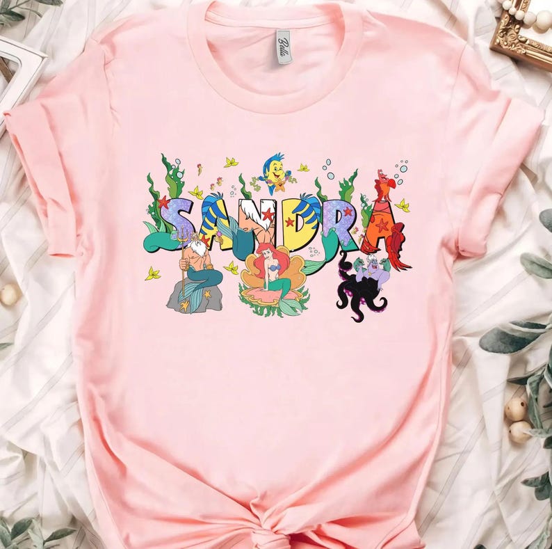 May include: A light pink t-shirt featuring the name "SANDRA" in colorful, whimsical lettering. Each letter is decorated with characters from a popular animated movie, including a mermaid, a crab, and a sea witch. The shirt is made of soft material.
