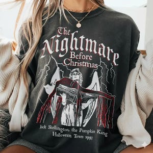 May include: Dark gray sweatshirt featuring the text "The Nightmare Before Christmas" in white and red, with an illustration of a character from the movie. The text "Jack Skellington, the Pumpkin King Halloween Town 1993" is also printed on the shirt.