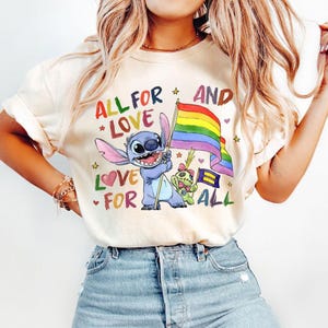 May include: Cream-colored t-shirt featuring a colorful graphic of Stitch holding a rainbow flag, with the text "ALL FOR LOVE AND LOVE FOR ALL". The design includes stars, hearts, and other characters.