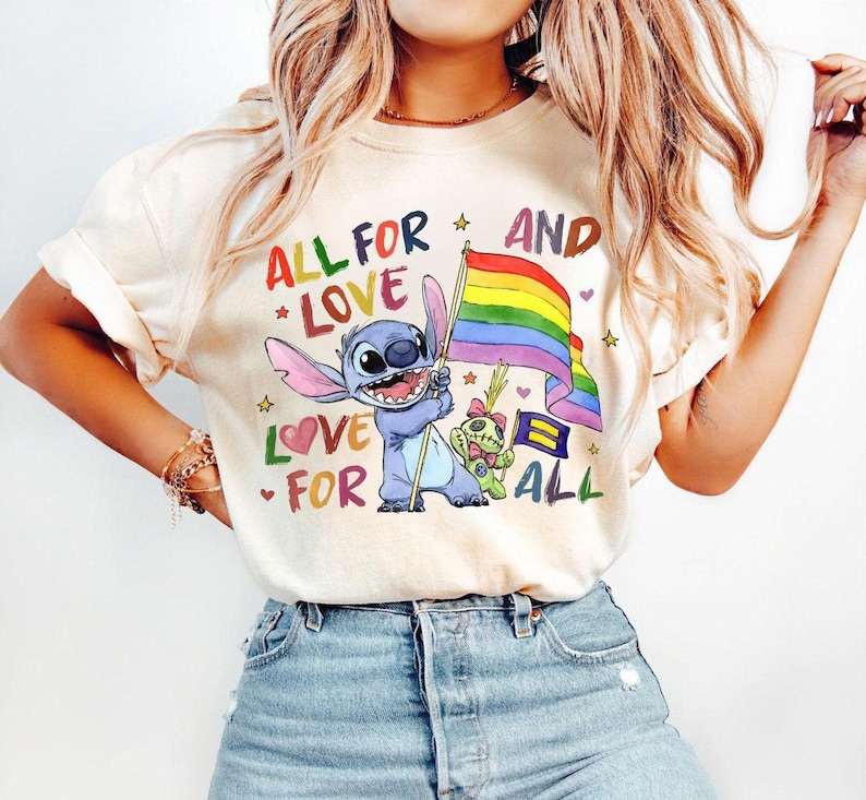 May include: Cream-colored t-shirt featuring a colorful graphic of Stitch holding a rainbow flag, with the text "ALL FOR LOVE AND LOVE FOR ALL". The design includes stars, hearts, and other characters.