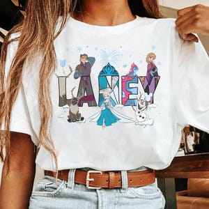 May include: White t-shirt with the word "LARKEY" in colourful, stylised letters. Each letter is decorated with characters from a popular animated film, including a reindeer, a snowman, and a princess.