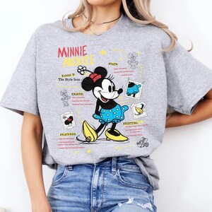 以下が含まれることがあります： ミニーマウスのグラフィックと赤い文字で「ミニーマウス」と書かれたライトグレーのTシャツ。ミニーは青い水玉模様のドレスと黄色の靴を履いています。シャツには追加のテキストとイラストも含まれています。