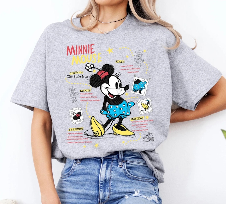以下が含まれることがあります： ミニーマウスのグラフィックと赤い文字で「ミニーマウス」と書かれたライトグレーのTシャツ。ミニーは青い水玉模様のドレスと黄色の靴を履いています。シャツには追加のテキストとイラストも含まれています。