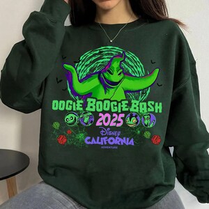 Op de afbeelding: Een donkergroen sweatshirt met lange mouwen en een afbeelding van het personage Oogie Boogie in felgroen. De tekst luidt Oogie Boogie Bash 2025 Disney California Adventure, vergezeld door kleine ronde karakterpictogrammen, dobbelstenen en spinnenwebben.