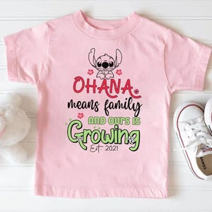 May include: A light pink t-shirt featuring a graphic of the character Stitch, with the words "Ohana means family and ours is Growing Est. 2021" in red and green. White sneakers are next to the shirt.