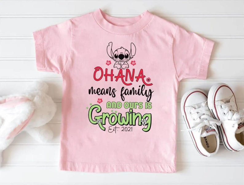 May include: A light pink t-shirt featuring a graphic of the character Stitch, with the words "Ohana means family and ours is Growing Est. 2021" in red and green. White sneakers are next to the shirt.
