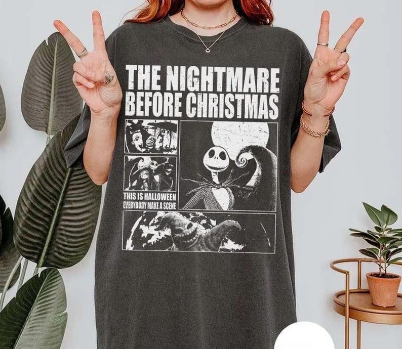 May include: A charcoal grey short-sleeve t-shirt featuring a white graphic print of The Nightmare Before Christmas. The design includes the movie title, scenes with Jack Skellington, and the text This is Halloween Everybody Make a Scene.
