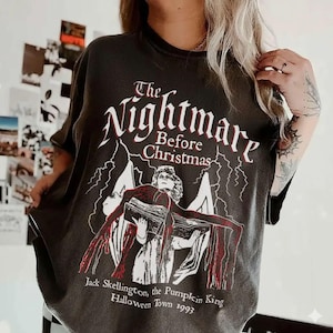 May include: Dark gray t-shirt featuring the text "The Nightmare Before Christmas" in white, with a graphic of a character holding a book. Below the graphic is the text "Jack Skellington, the Pumpkin King, Halloween Town 1993."