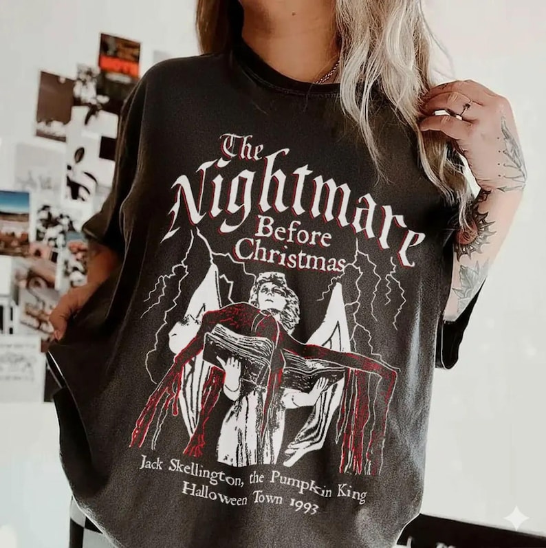 May include: Dark gray t-shirt featuring the text "The Nightmare Before Christmas" in white, with a graphic of a character holding a book. Below the graphic is the text "Jack Skellington, the Pumpkin King, Halloween Town 1993."
