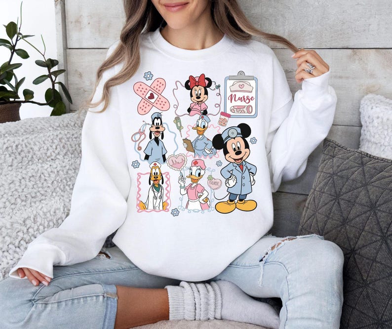 Puede incluir: Sudadera blanca con personajes de dibujos animados vestidos como profesionales m&eacute;dicos. Minnie Mouse, Donald Duck, Mickey Mouse y otros est&aacute;n representados con batas y batas de laboratorio, con s&iacute;mbolos m&eacute;dicos como estetoscopios y vendas.