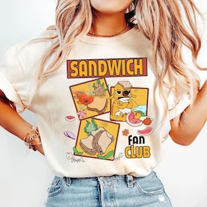 May include: Cream-colored t-shirt featuring the words "SANDWICH FAN CLUB" in a brown and yellow design. The shirt has three cartoon sandwich illustrations with various toppings and a fish, dog, and sandwich.