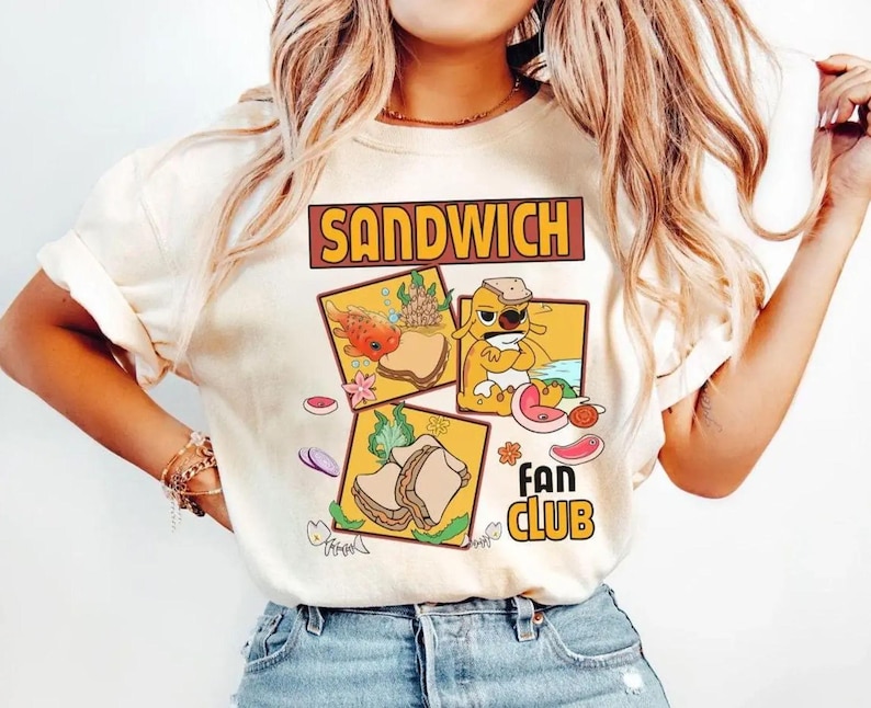 May include: Cream-colored t-shirt featuring the words "SANDWICH FAN CLUB" in a brown and yellow design. The shirt has three cartoon sandwich illustrations with various toppings and a fish, dog, and sandwich.