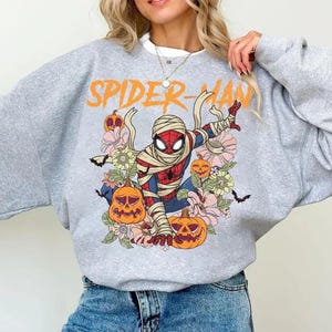 K&ouml;nnte beinhalten: Graues Sweatshirt mit einem Halloween-Thema Spider-Man-Design. Die Grafik zeigt Spider-Man in Bandagen, K&uuml;rbissen, Blumen und dem orangefarbenen Text "SPIDER-MAN".