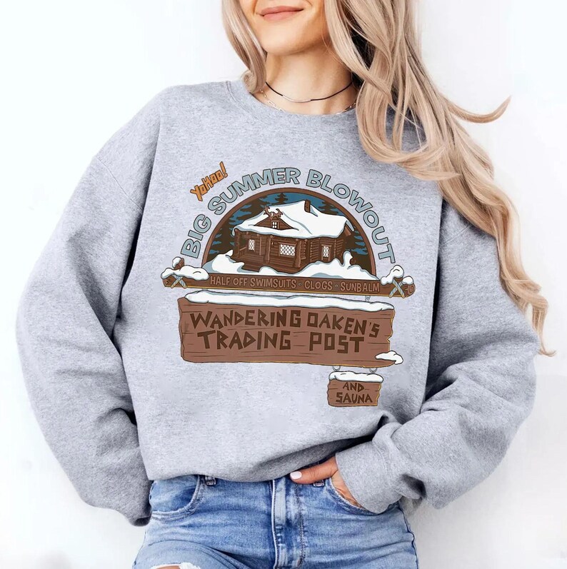 Op de afbeelding: Een lichtgrijs sweatshirt met een grafische opdruk met de tekst "BIG SUMMER BLOWOUT" boven een hutillustratie. Daaronder staat een bord met de tekst "WANDERING OAKEN'S TRADING POST AND SAUNA".