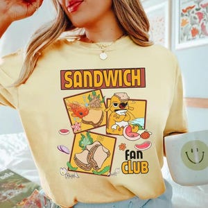 May include: Yellow t-shirt with the word "SANDWICH" in a brown rectangular box. The shirt features a graphic of a sandwich and the words "FAN CLUB" in a yellow box with a flower.