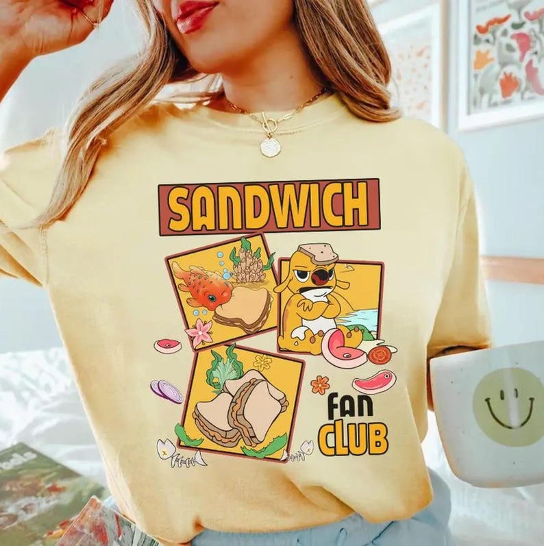 May include: Yellow t-shirt with the word "SANDWICH" in a brown rectangular box. The shirt features a graphic of a sandwich and the words "FAN CLUB" in a yellow box with a flower.